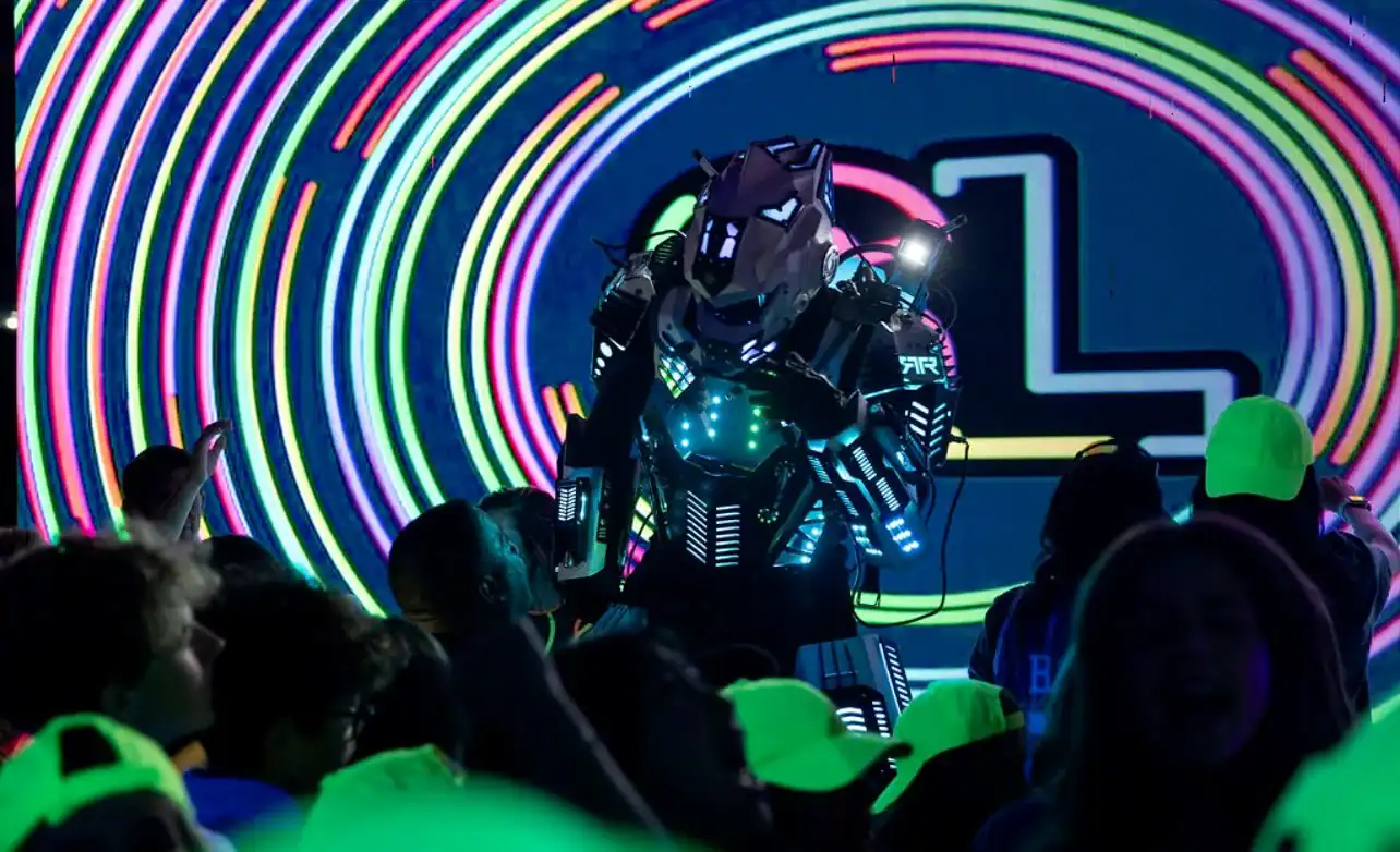 A person in a glowing, futuristic robot costume stands amid a crowd at a neon-lit event, designed by an expert event planner Nassau County, with colorful circular light patterns displayed on a large screen in the background.
