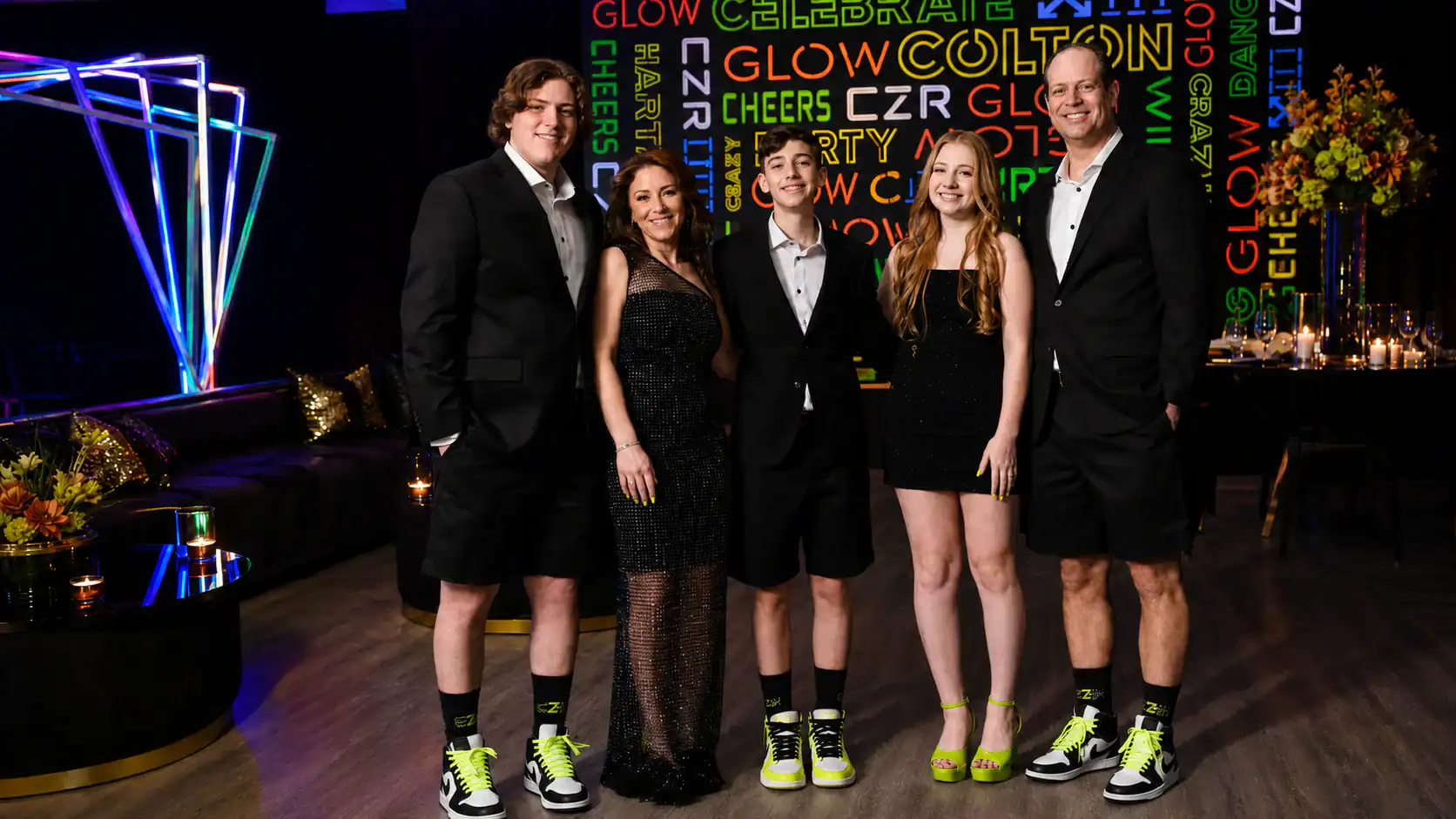 Five people dressed in black formal wear and neon-accented shoes and socks pose together at a party. A neon-lit, festive backdrop with words like "celebrate" and "glow" is visible behind them.